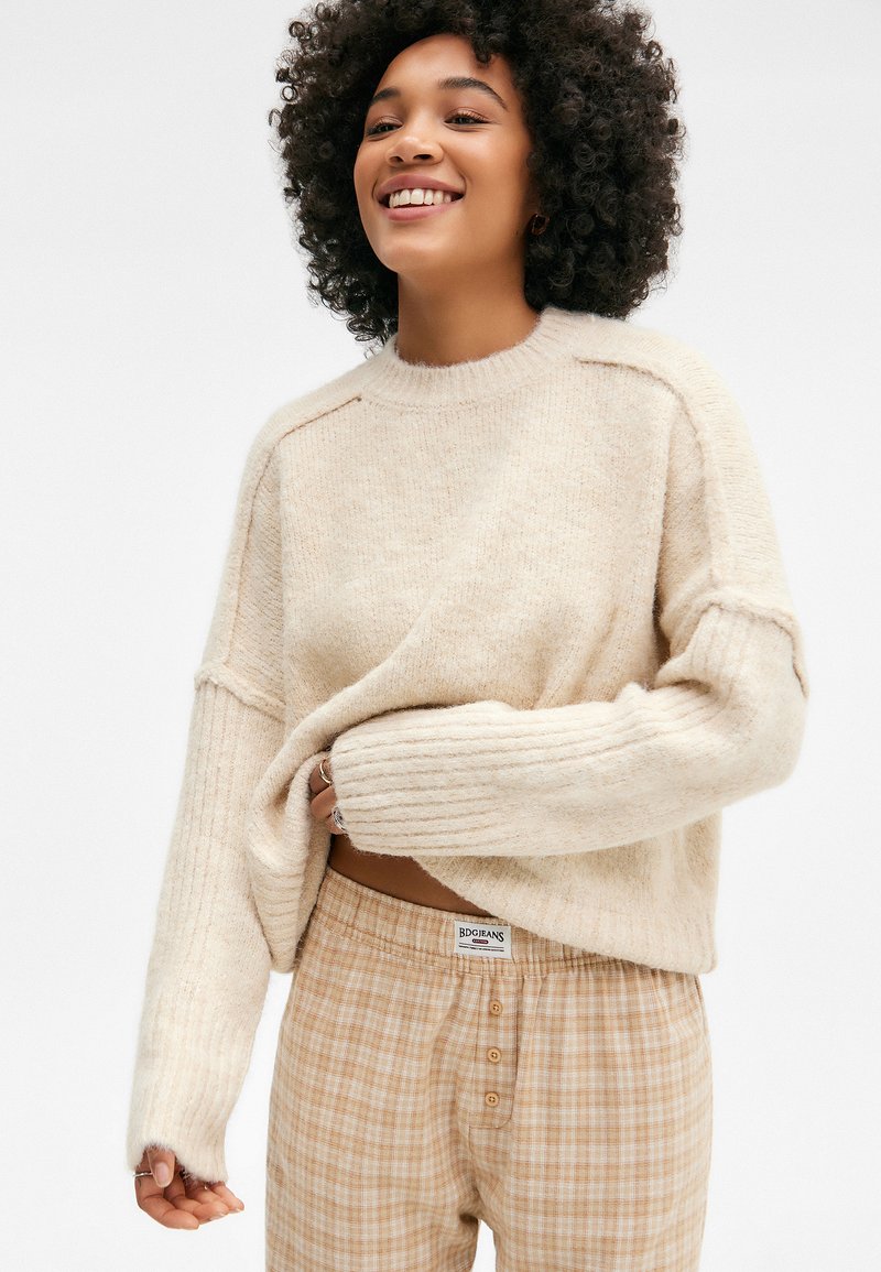 Hellbeige, grob gestrickter Pullover mit Rippbündchen und Rundhalsausschnitt, kombiniert mit taubrechten, karrierten, knopfgestützten Hosen. Weiche Textur im gesamten Outfit.