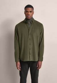 Grüne Hemdbluse aus weichem Stoff mit einer Tasche. Lange Ärmel, gerader Schnitt und schwarze Knöpfe. Über einem dunklen Rollkragenpullover getragen.