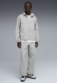 Man in een lichtgrijze sportjack en -broek met witte piping, witte sneakers, zwarte zonnebril en zilveren kettingen, staand op een grijze achtergrond.