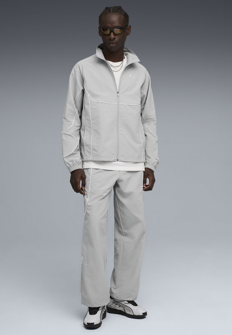 Man in een lichtgrijze sportjack en -broek met witte piping, witte sneakers, zwarte zonnebril en zilveren kettingen, staand op een grijze achtergrond.