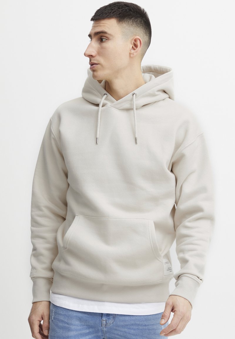 Sweat à capuche beige en tissu doux, doté d'une poche avant, de cordons de serrage et de poignets côtelés. Design simple sans motifs.