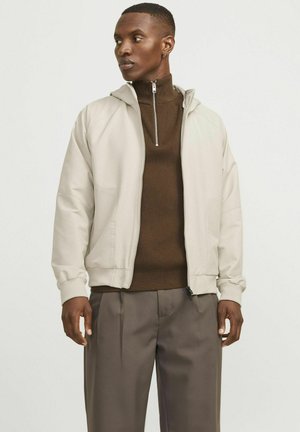 Veste zippée beige clair avec capuche, en tissu texturé et poignets côtelés, portée sur un haut marron à col montant et un pantalon gris.