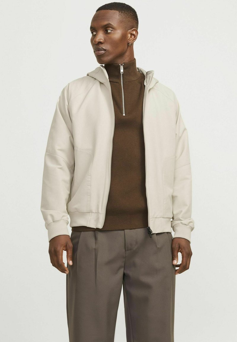 Veste zippée beige clair avec capuche, en tissu texturé et poignets côtelés, portée sur un haut marron à col montant et un pantalon gris.