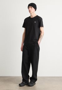 Svart bomull t-skjorte med en liten logo, kombinert med løse svarte bukser og svarte sneakers. Modellen har også på seg en svart strikket beanie.