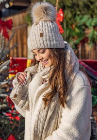 Gorro de punto beige con un pom-pom esponjoso, combinado con una bufanda texturizada beige y una chaqueta cómoda de color crema. Presenta un pequeño acento de logo.