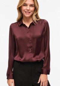 Blouse en satin bordeaux à manches longues, fermeture boutonnée à l'avant, et col classique. Texture lisse avec un éclat subtil et poignets.