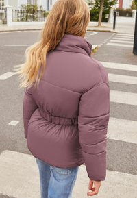 Pufjakas pelēkā rozā krāsā ar jostu viduklī, lielu apkakli un quiltētu tekstūru. Apvienots ar zilu džinsu pie gājēju pārejas.
