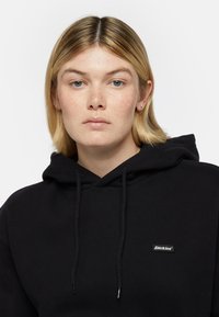 Felpa nera con cappuccio, con tasca frontale, cordini e una piccola etichetta con il logo "Dickies" sul petto. Tessuto morbido, vestibilità comoda.