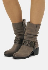 Bottines taupe à la cheville avec un design ample, dot�ées d'une bande décorative avec des accents triangulaires, d'une fermeture éclair et d'un talon épais.