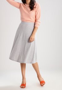Donna in un maglione pesca, una gonna midi grigia chiara a forma di A e scarpe con apertura arancione brillante, in posa contro uno sfondo leggero e uniforme.