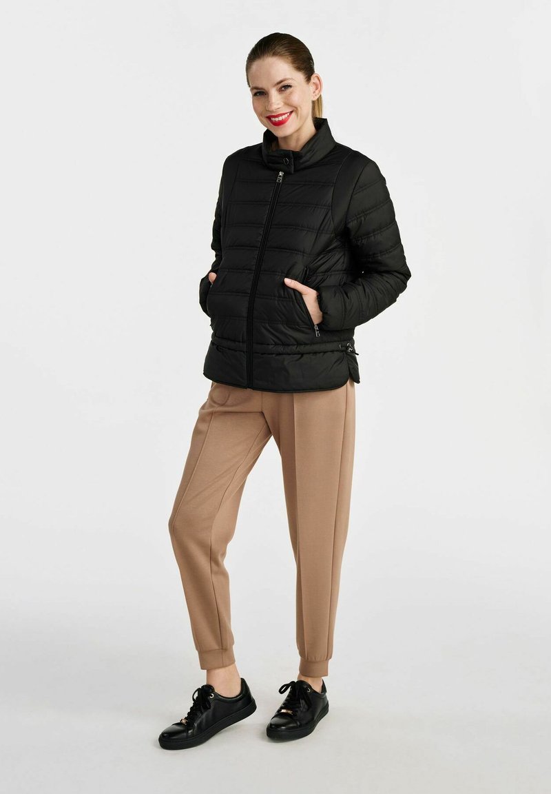 Czarna kurtka puchowa z poziomym wzorem przeszyć, wysokim kołnierzem, zapięciem na zamek oraz dwoma bocznymi kieszeniami, w połączeniu z beżowymi spodniami i czarnymi sneakersami.