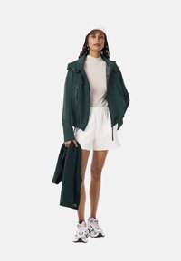 Donkergroene jas met een capuchon, beige geribde trui, witte shorts, houdt een donkergroen gebreid sweatervest vast, draagt witte sneakers en een witte pet.