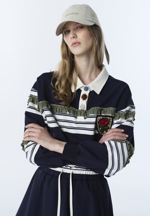 Vrouw draagt een marineblauw gestreept topje met olijfgroene ruches, een rozenpatch, een beige kraag met decoratieve knopen, een marineblauwe rok en een beige honkbalpet.