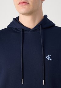 Felpa con cappuccio blu navy con coulisse, tessuto morbido e logo ricamato azzurro sul petto. Texture liscia e design casual.