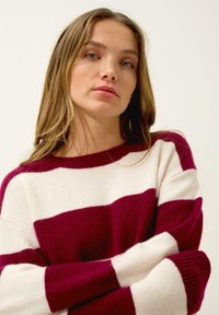 Pull en tricot rayé bordeaux et crème avec une coupe décontractée, un col rond et des poignets côtelés. Tissu doux et texturé.