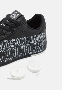 Baskets noires avec texte blanc « Versace Jeans Couture » sur le côté, lacets noirs et deux lacets blancs roulés à côté, sur fond blanc.