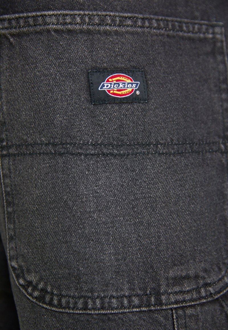 Zwart denim stof heeft een opvallend Dickies-logo op de rechterzak, met zichtbare stiksels en gestructureerde oppervlaktedetails.