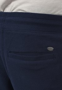Pantalon bleu marine en tissu texturé et côtelé. Doté d'une poche arrière avec une étiquette ovale en métal argenté. Ceinture visible juste au-dessus du pantalon.