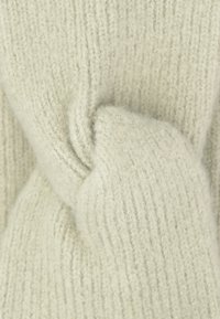 Jasno beżowy sweter dziany o miękkiej, teksturowanej powierzchni. Tkanina ma prążkowane wzory oraz skręcony detal z przodu.