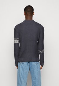 Harmaa neulepusero, jossa on resorivetoketju helman ja hihansuiden kohdalla ja kuvioidut yksityiskohdat hihoissa. Yhdistetty vaaleansinisiin, väljämallisiin farkkuihin.