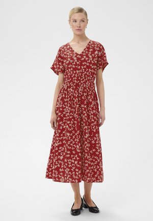 Žena plave kose u crvenoj midi haljini s kratkim rukavima i cvjetnim uzorkom te crnim cipelama s niskom petom stoji na jednoličnoj pozadini.