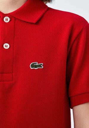 Polo rouge en tissu texturé avec un col, doté de deux boutons blancs et d'un logo de crocodile vert brodé sur la poitrine gauche.