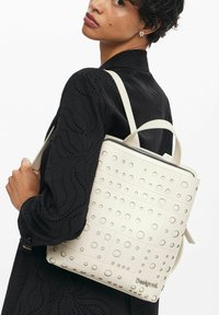 Mochila blanca texturizada con patrones circulares, diseño cuadrado y un cierre superior de cremallera. Correas ajustables y un asa resistente para su transporte.