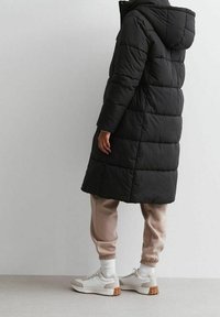 Musta, tilkkuompelemalla tikattu pitkä toppatakki hupulla. Löysä istuvuus, vaakasuorat tikit ja korkea kaulus. Yhdistetty vaaleanbeigeihin collegehousuihin ja lenkkareihin.