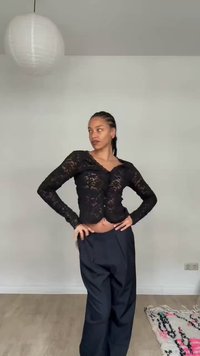 Femme aux cheveux tressés posant avec les mains sur les hanches, portant un haut noir en dentelle à manches longues et un pantalon noir ample dans une pièce minimaliste avec une lumière suspendue.