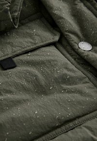 Tessuto verde scuro, resistente all'acqua, con gocce visibili. Presenta una superficie testurizzata e un bottone a scatto metallico. Accentuato da un cappio nero.
