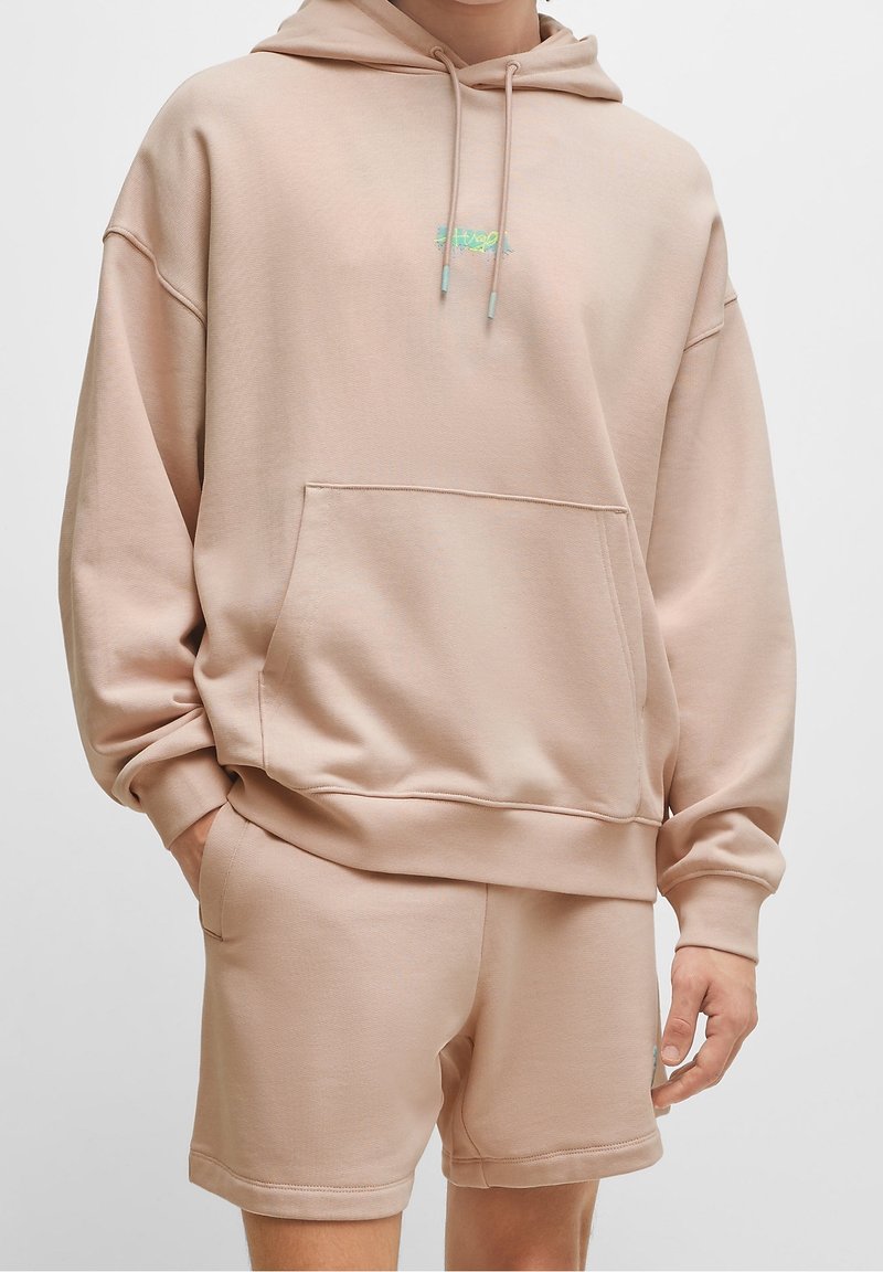 Homme portant un sweat à capuche beige oversize avec poche avant et un short assorti, debout devant un fond blanc uni.