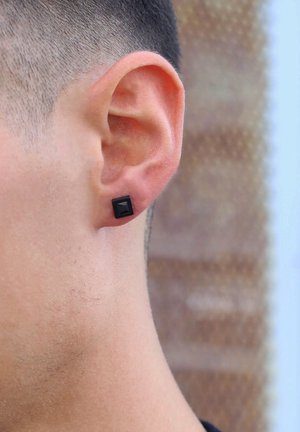Square black stud earring with a raised pyramid detail, set on the lower lobe of the ear against a neutral skin tone.