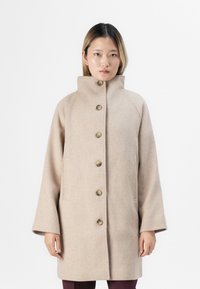 Personne portant un manteau beige boutonné avec un col haut et des manches larges, se tenant contre un fond blanc uni.