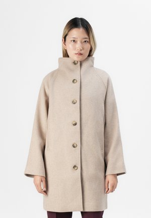 Personne portant un manteau beige boutonné avec un col haut et des manches larges, se tenant contre un fond blanc uni.