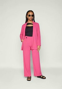 Fuchsia pink suit with a tailored blazer and wide-leg trousers. Black top with a cutout detail. Model wearing dark sunglasses and sandals.