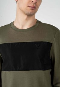 Maglione verde oliva con un pannello in nylon nero sul petto. Caratterizzato da un collo a giro e un logo triangolare nella parte superiore sinistra del petto.