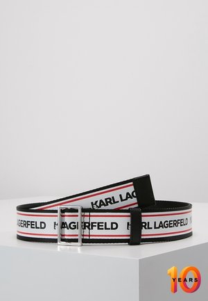 Biały pas materiałowy z czarnymi brzegami, czerwoną paską, powtarzającym się napisem „KARL LAGERFELD” oraz srebrną, prostokątną klamrą, wyświetlany na białej powierzchni.