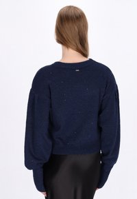 Navyblauwe, cropped gebreide trui met pofmouwen en een subtiele glans. Heeft een geribde zoom en een labeldetail aan de achterkant. Gladde textuur.