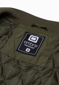 Interno di una giacca trapuntata verde con un'etichetta nera con la scritta "ombre menswear M" attaccata al collo, dotata di un colletto a coste e una texture liscia.