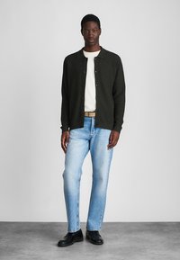 Tmavě zelený cardigan s knoflíky přes bílé tričko, spárovaný s světle modrými džíny a černými botami. Jednoduchý design a texturovaná látka.