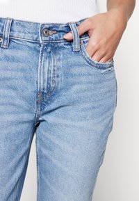 Jeans de mezclilla azul claro con cintura alta, costuras visibles, bolsillos frontales y cierre de botón metálico. La tela tiene una textura ligeramente deslavada.