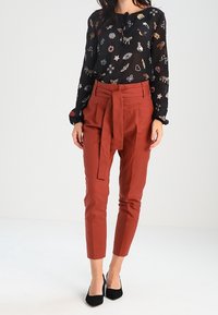 Schwarze durchsichtige Bluse mit Tier- und Blumenmustern, kombiniert mit hochtaillierten Hosen in Rostfarbe mit Bindeband und schwarzen Absätzen.