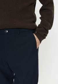 Tamsiai mėlyni audinio kelnės su priekiu kišene ir sagos užsegimu. Pateiktos kartu su rudu tekstūruotu megztiniu.