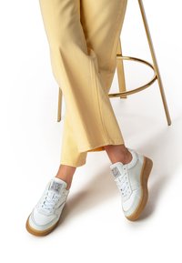 Sneakers bianche in pelle con suole in gomma, caratterizzate da un design texturizzato, lacci e logo sulla linguetta, abbinate a pantaloni giallo chiaro a gamba larga.