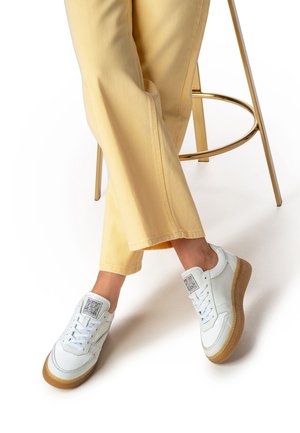 Baskets blanches en cuir avec semelles en caoutchouc, présentant un design texturé, des lacets et un logo sur la languette, associées à un pantalon large jaune clair.