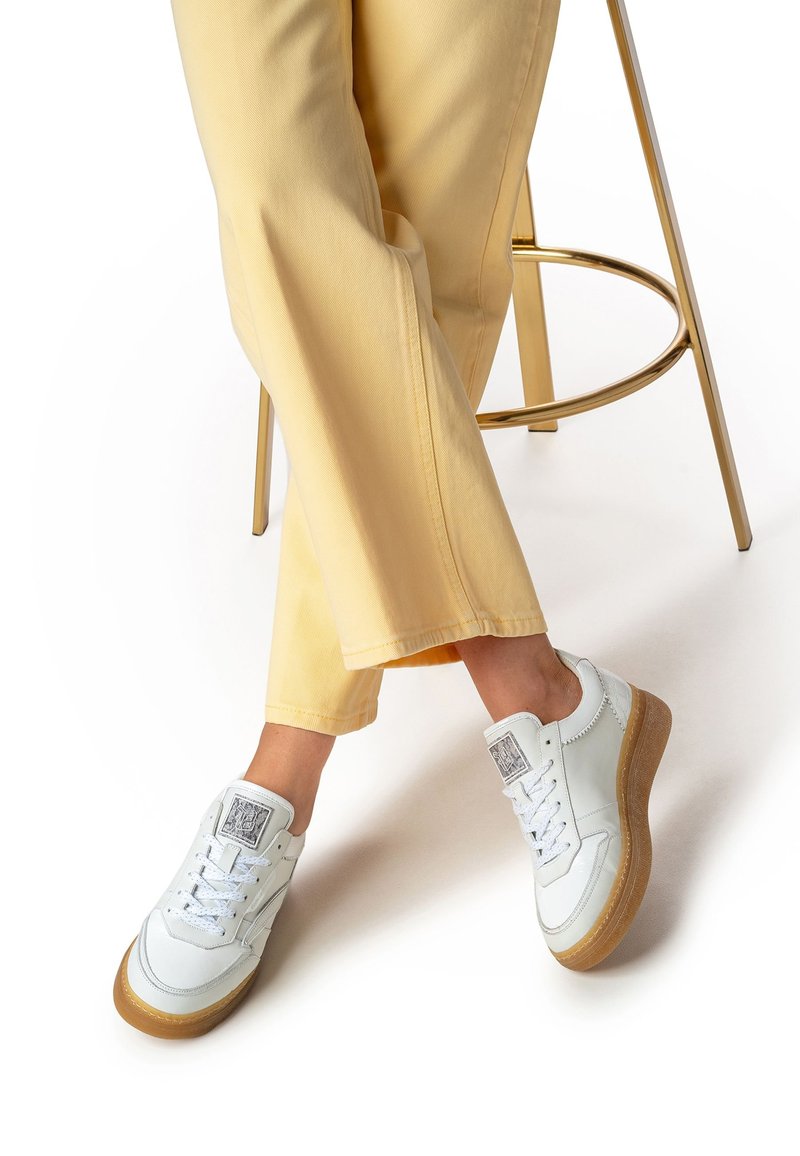 Sneakers bianche in pelle con suole in gomma, caratterizzate da un design texturizzato, lacci e logo sulla linguetta, abbinate a pantaloni giallo chiaro a gamba larga.