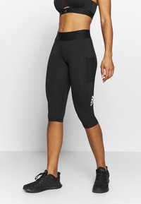 Kvinna i svarta Adidas cropped leggings med sidoficka och svarta träningsskor mot vit bakgrund.