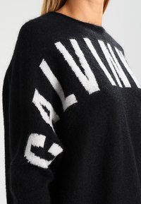 Pull noir avec logo blanc, conçu dans un matériau doux, présentant une coupe décontractée et un col rond. Remarquable par son design surdimensionné et son contraste de couleurs.