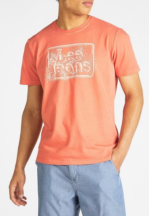 Camiseta coral de manga corta de algodón, con un logo blanco de "Lee Jeans" en un diseño rectangular, combinada con pantalones rayados azul claro.