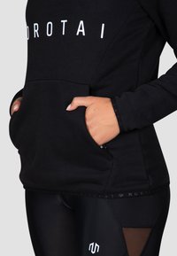 Schwarzer Hoodie mit Kängurutasche, ausgestattet mit auffälligen weißen Buchstaben und gerippten Bündchen, kombiniert mit schwarzen Leggings mit Mesh-Akzenten.